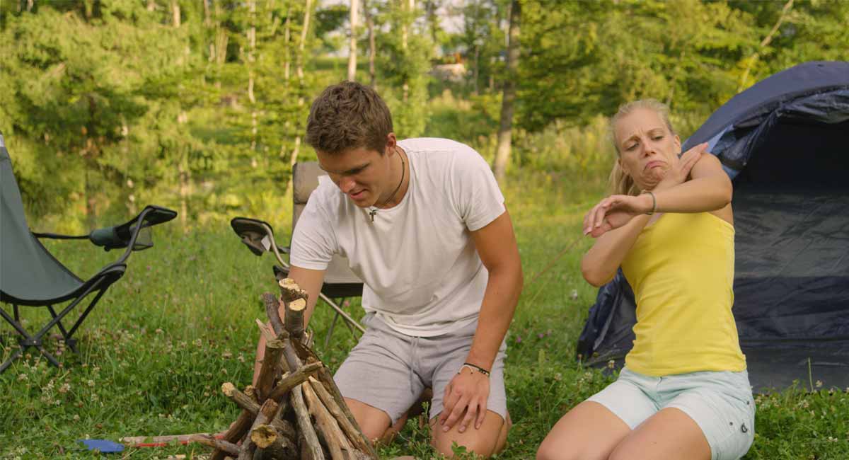 man building fire and woman swatting off mosquitos